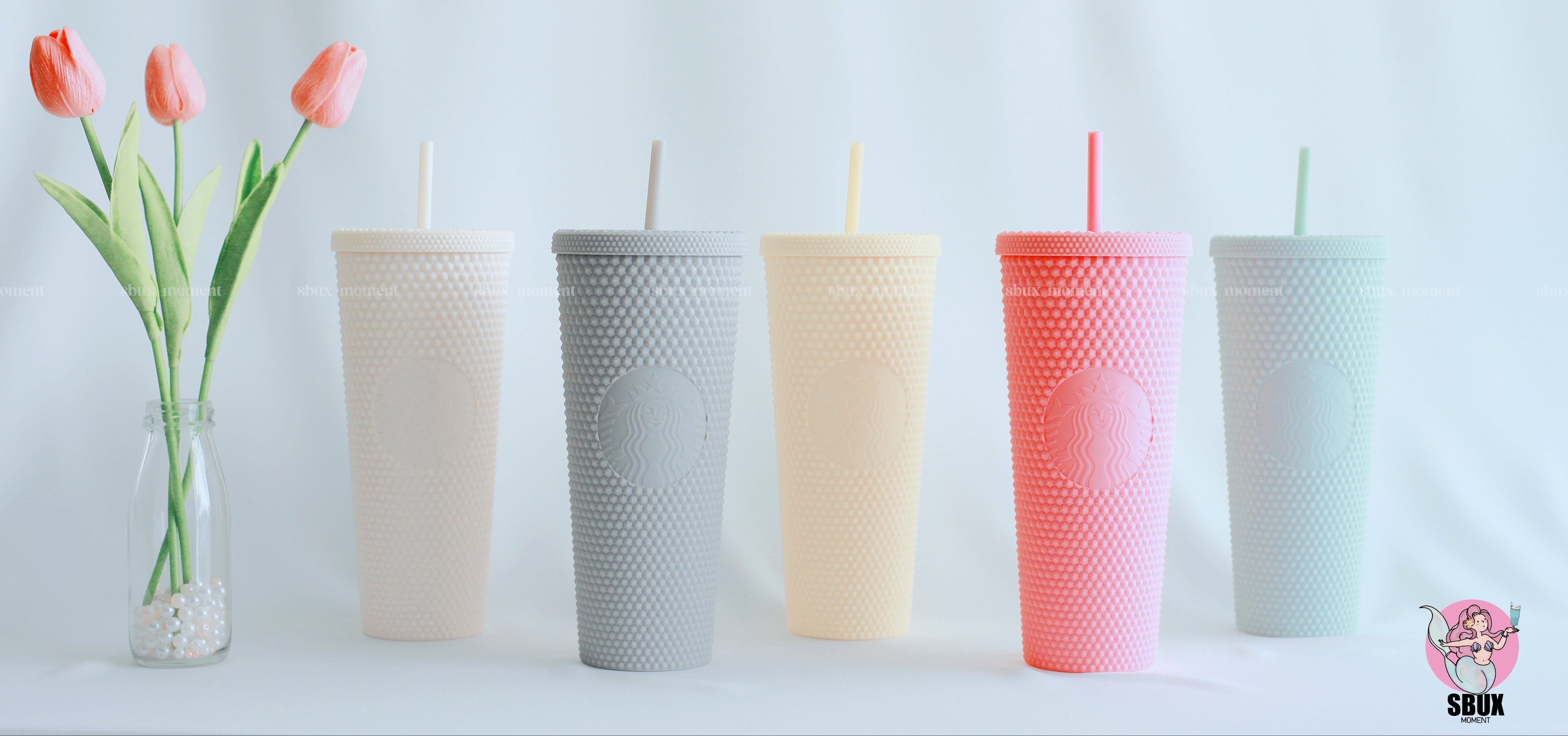 Five colorful tumblers with straws on a light blue background, accompanied by a vase of tulips.